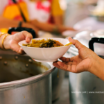 Sobre um panelão de sopa, uma mão entrega uma tigela de sopa para outra mão que a recebe