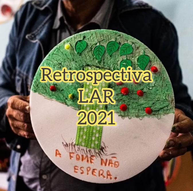 Uma par de mãos segura uma pintura em tela redonda com uma árvore pintada, onde foram anexadas folhas verdes recortadas de papel e bolinhas amarelas e vermelhas. a frase escriva em giz de cera "A Fome Não Espera"