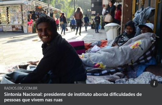 Um homem em situação de rua sorri para a camera sentado ao sol onde 2 outros moradores dormem enrolados em suas cobertas.